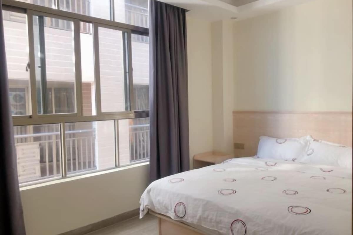 Une chambre d’hôtel avec une grande fenêtre, un lit recouvert d’une courtepointe blanche, une table de chevet et une tête de lit.