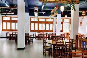 A spacious dining area with multiple wooden tables and chairs, large windows, and hanging plants.