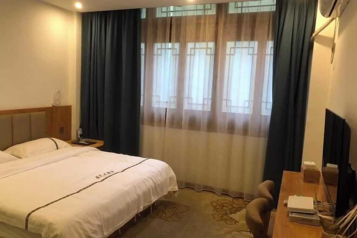 A hotel room with a bed, a desk, and a chair. There is a window with curtains and a view of the outside.