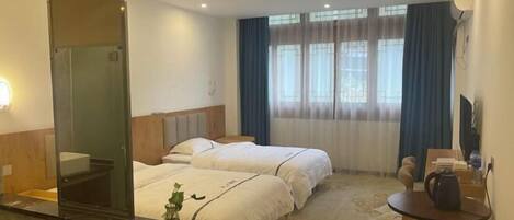 A hotel room with two beds, a small table, a chair, a desk, a mirror, and a window with curtains.