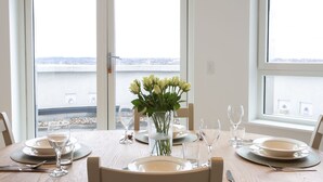 Een eettafel gedekt met borden, glazen en een bloevas, met uitzicht op een waterpartij door grote ramen.