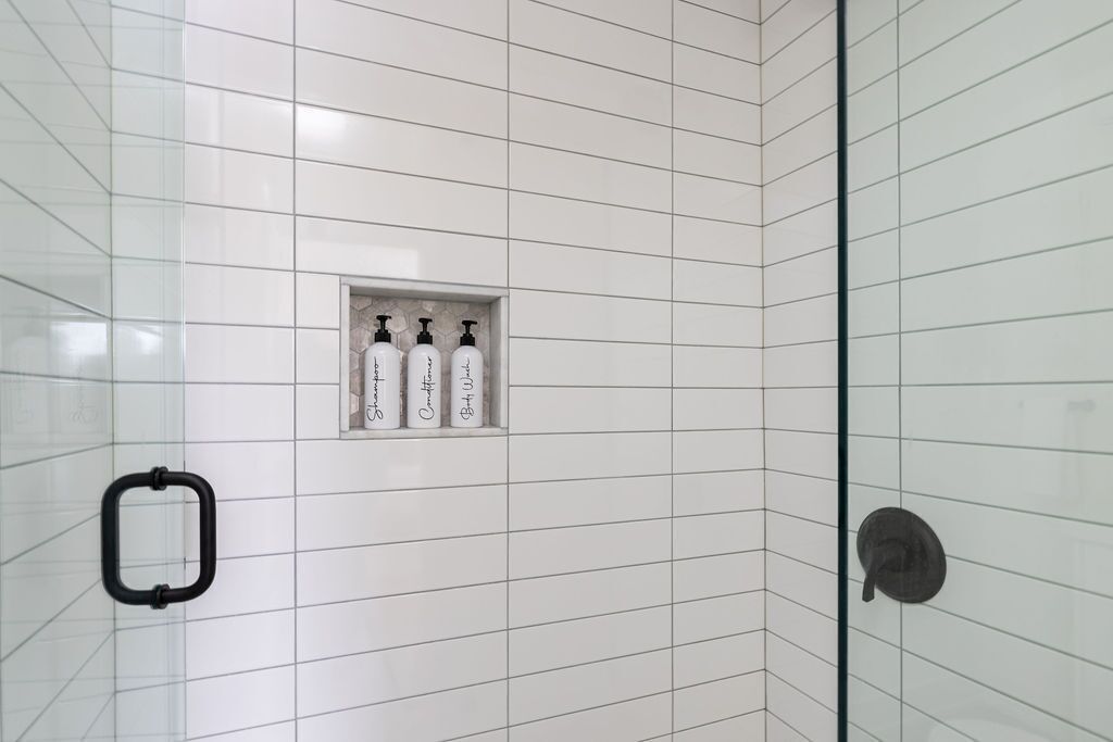 A shower area with white tiles, a glass door, and a shelf with three bottles.