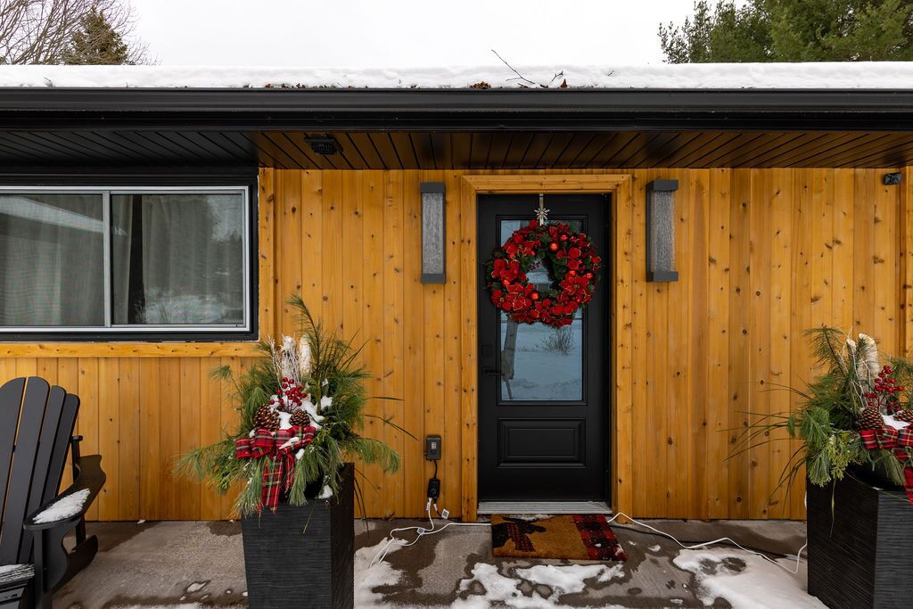 Een houten huis met een zwarte deur, een rode krans en twee potten met groen en dennenappels.