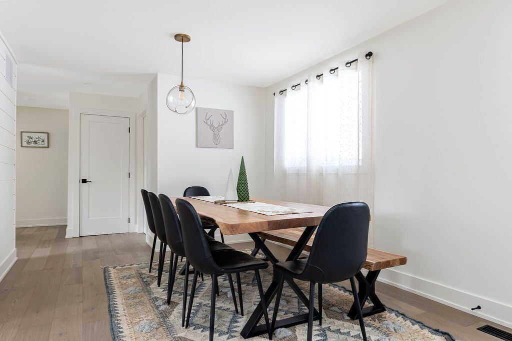 Een eetgedeelte met een houten tafel, zwarte stoelen, een kamerkleed met patroon, een hanglamp en een raam met dunne gordijnen.