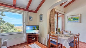 Uma área de refeições com mesa posta para dois, uma televisão sobre um suporte e uma janela com vista para o mar.