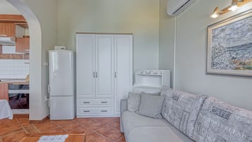 Un espacio habitable compacto con kitchenette, un armario blanco, un sofá gris con cojines estampados y un cuadro en la pared.