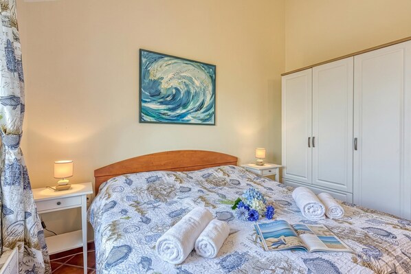 Un lit bien fait, recouvert d’une couvre-lit à motifs, un tableau au mur, une armoire blanche et une table de chevet avec une lampe.