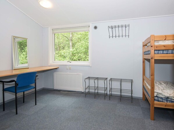 Quarto com cama de solteiro, escrivaninha, cadeira, radiador e janela com vista para árvores.