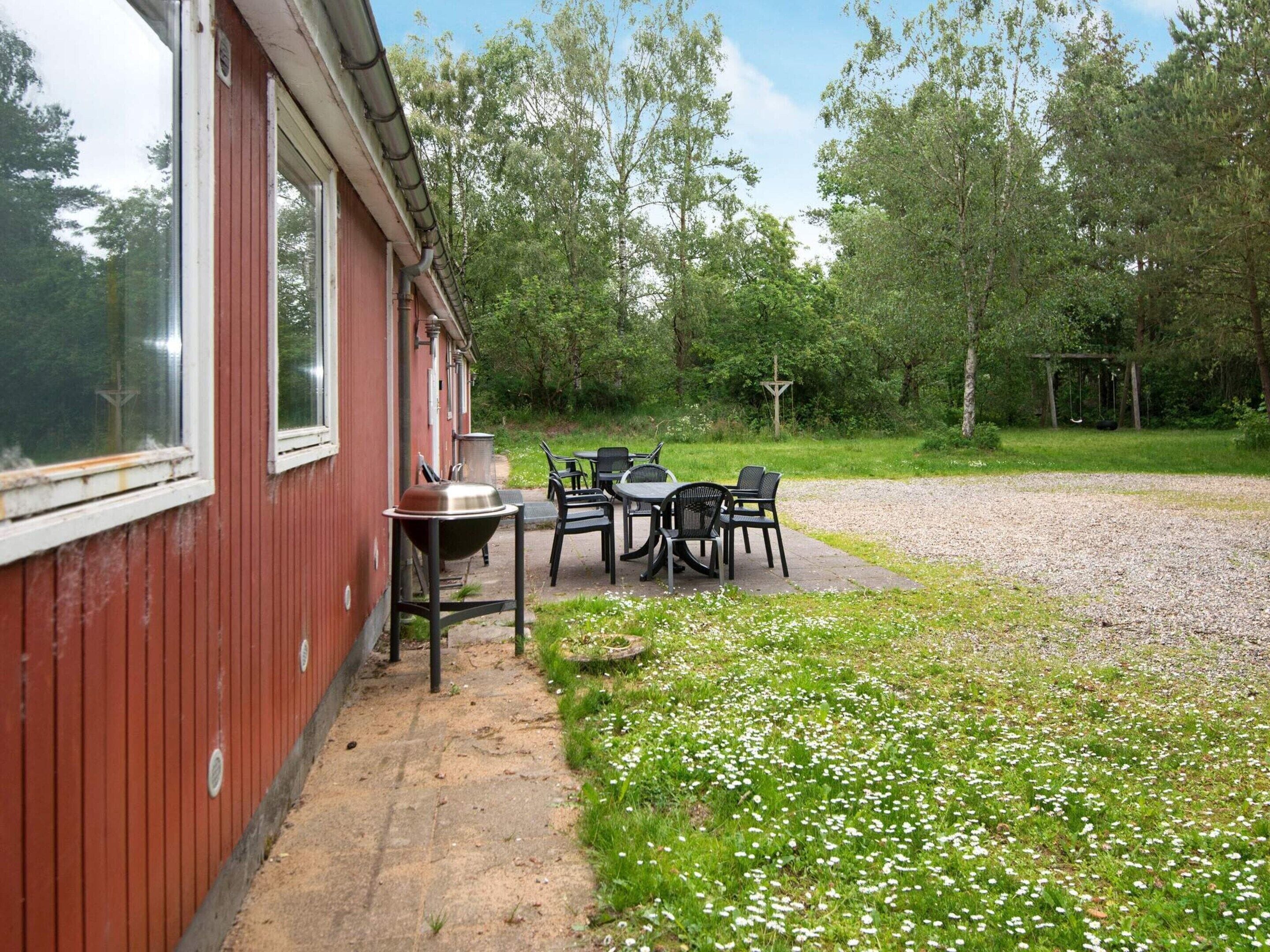 A red building with a barbecue grill outside, surrounded by a gravel area and a grassy field with white flowers.