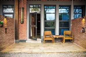 Une façade en briques agrémentée de grandes fenêtres, d’une porte et de deux fauteuils en osier sur une terrasse carrelée.