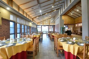 Un comedor amplio con varias mesas dispuestas para comidas, techos altos con vigas a la vista y amplias ventanas que dejan entrar luz natural.