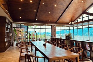 Une salle à manger spacieuse avec une longue table en bois et des chaises, de grandes fenêtres offrant une vue dégagée et une bibliothèque remplie de livres.