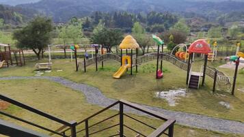 Un parque infantil con toboganes y estructuras para escalar, rodeado de césped y árboles, con montañas al fondo.