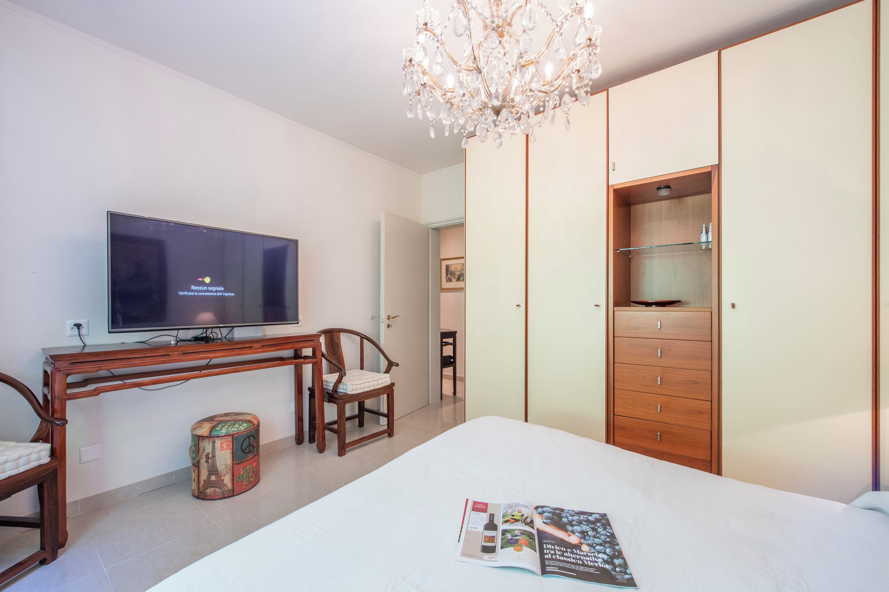 A bedroom with a bed, a TV, a wooden desk, a chair, a small round table, and a wardrobe.