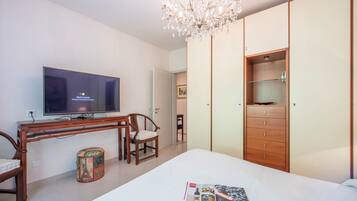 A bedroom with a bed, a TV, a wooden desk, a chair, a small round table, and a wardrobe.