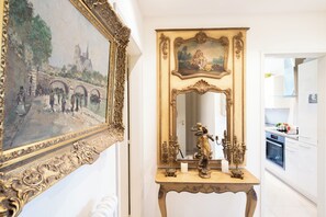 Ein Zimmer mit einem großen, verzierten Spiegel, einem Gemälde einer Brücke und einem kleinen Tisch mit Kerze und Statue.