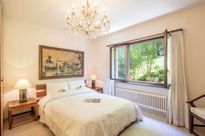 A bedroom with a large bed, a chandelier, a window with curtains, a radiator, and a painting on the wall. - Queen Lake View Apartment With Terrace (Tresa)