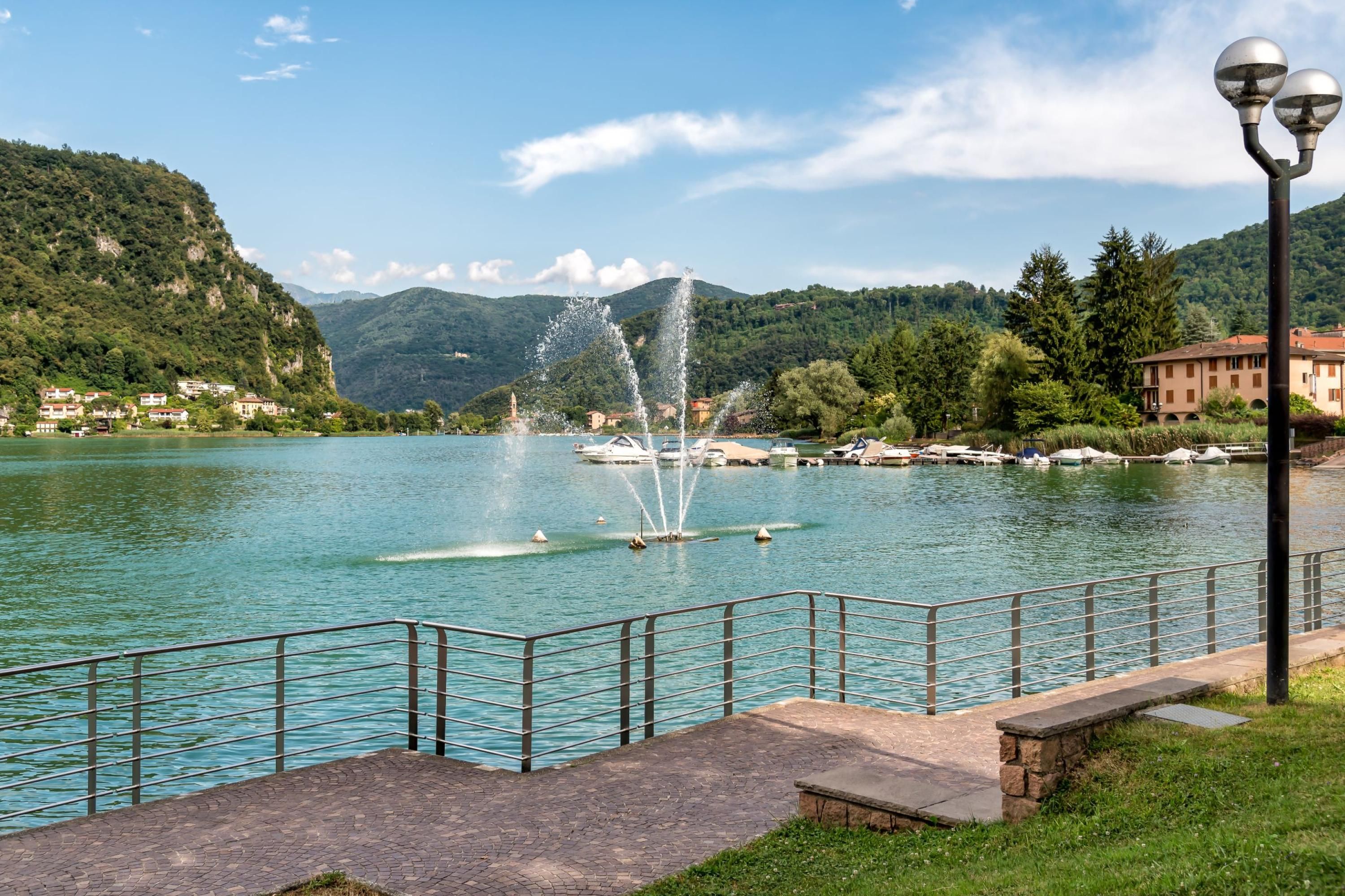 A waterfront promenade with a fountain, a stone bench, and a metal railing.