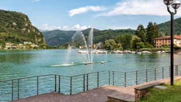 A waterfront promenade with a fountain, a stone bench, and a metal railing.