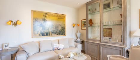 Uma sala de estar com um sofá bege, uma mesa de centro com objetos decorativos, um aparador de vidro com copos e um quadro grande na parede.