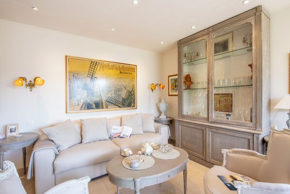 Un salon avec un canapé beige, une table basse agrémentée d’objets décoratifs, une vitrine contenant de la verrerie, et un grand tableau encadré accroché au mur.