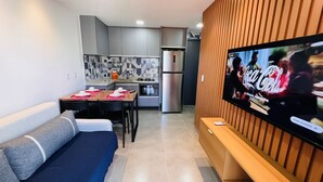 Una sala de estar moderna con un sofá, una mesa de comedor con platos y tazas, una zona de cocina con refrigerador y microondas, y un televisor mostrando un anuncio.
