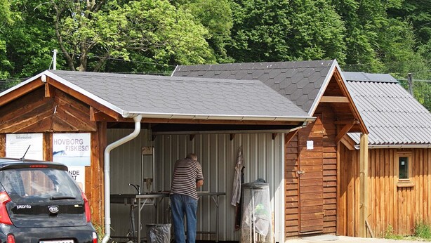 A small wooden building with a metal roof, a man standing at a sink, and a car parked in front. - 5 Person Holiday Home in Hovborg (Hovborg)