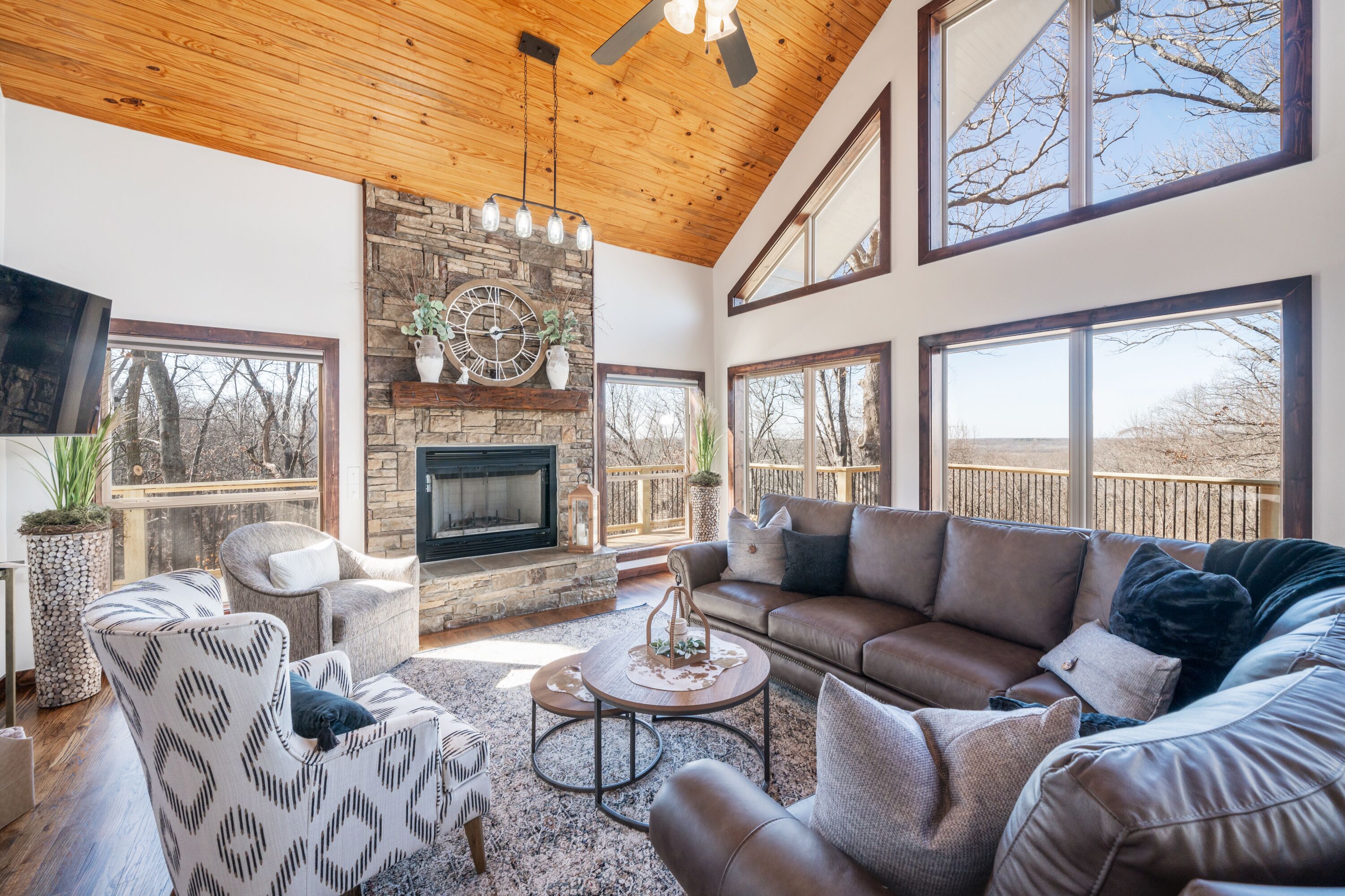 A spacious living room with a stone fireplace, large windows, and a high ceiling.