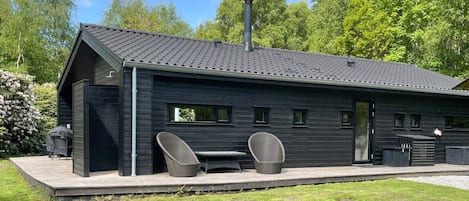 A modern black building with a sloped roof, a covered patio with two chairs and a table, and a chimney.