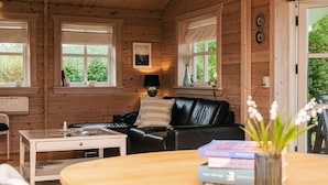 A cozy cabin interior with a black leather couch, a wooden dining table, and a small white table. There are two windows with white blinds, a lamp on the wall, and a door leading outside. - 4 Person Holiday Home in Ega (Aarhus)