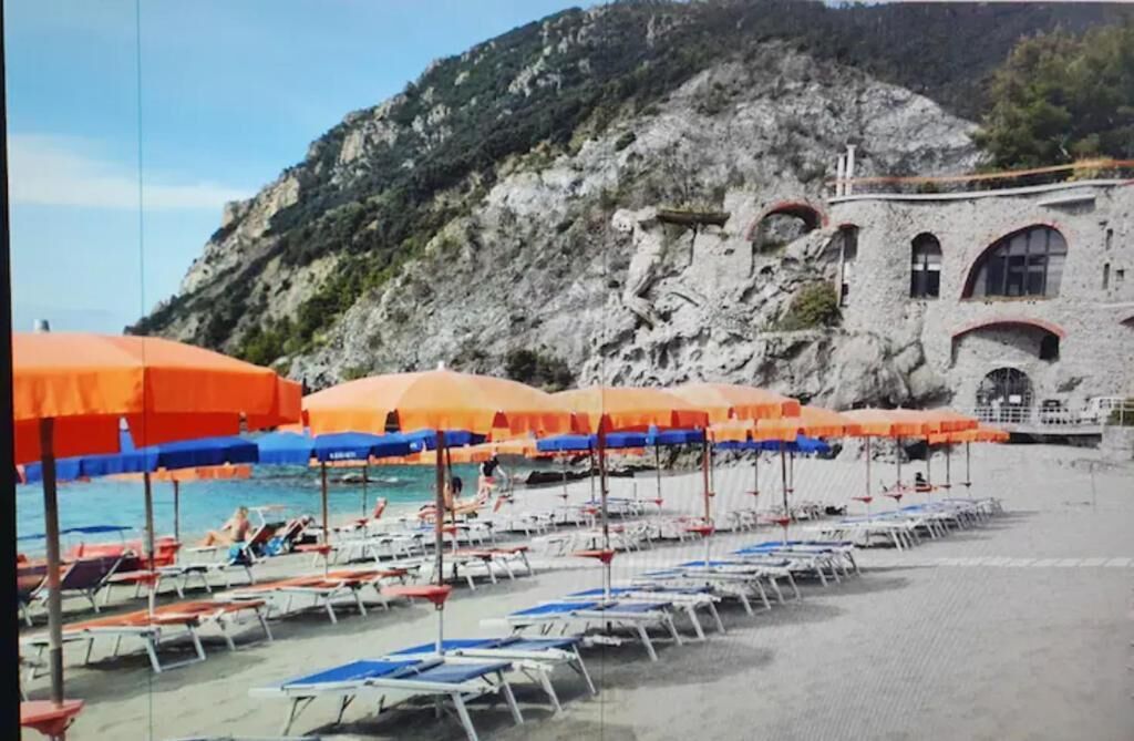 A beach with rows of sun loungers and orange umbrellas, a rocky mountain in the background, and a building with arched windows.