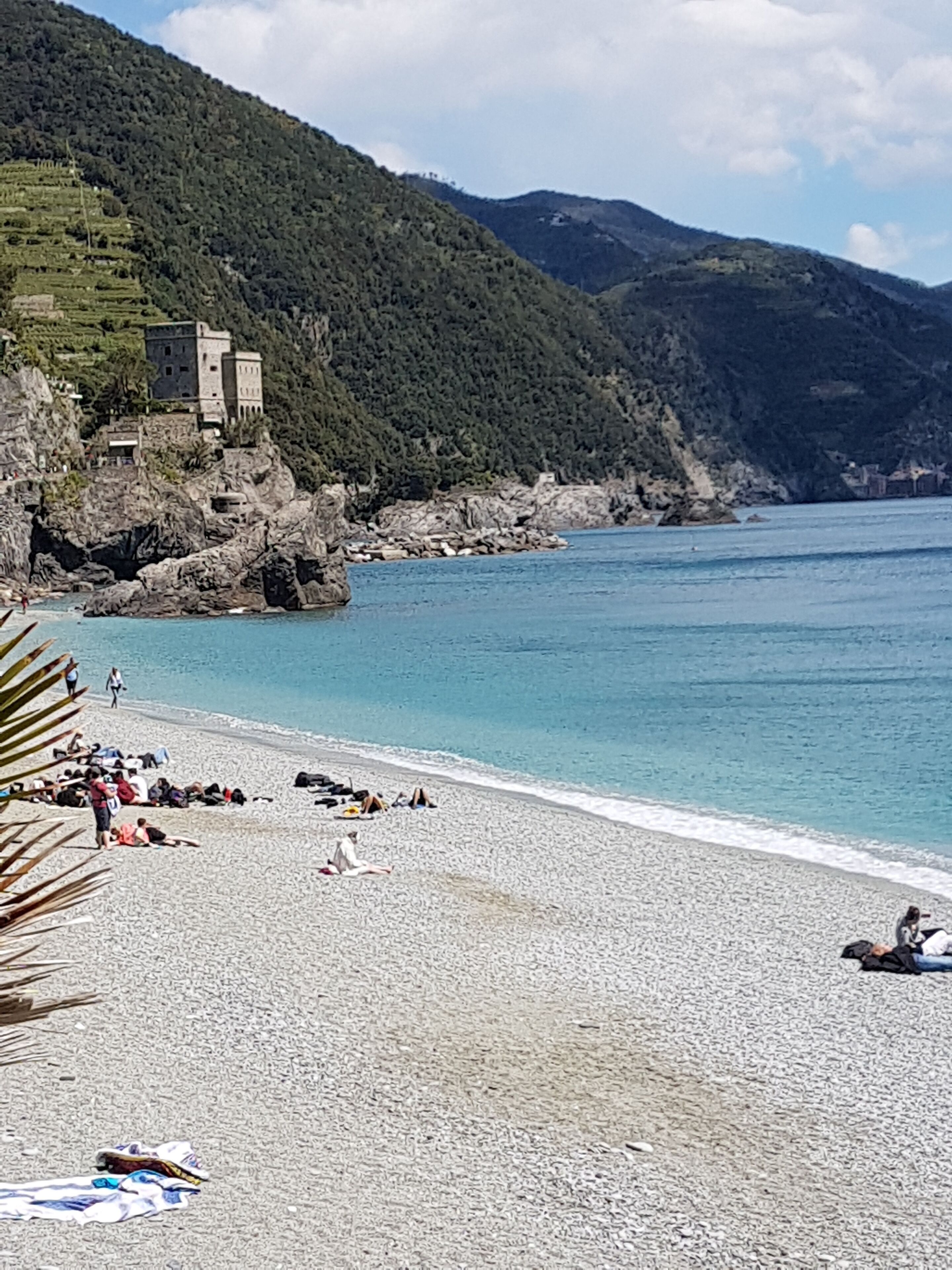 Ein Kiesstrand mit einem Schloss auf einem Hügel, klares blaues Meer und Berge im Hintergrund.