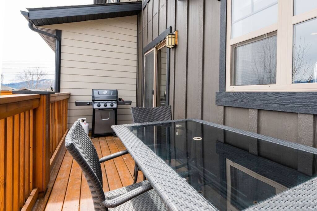 An outdoor patio with a glass-top table and wicker chairs, a barbecue grill, and a wooden deck.