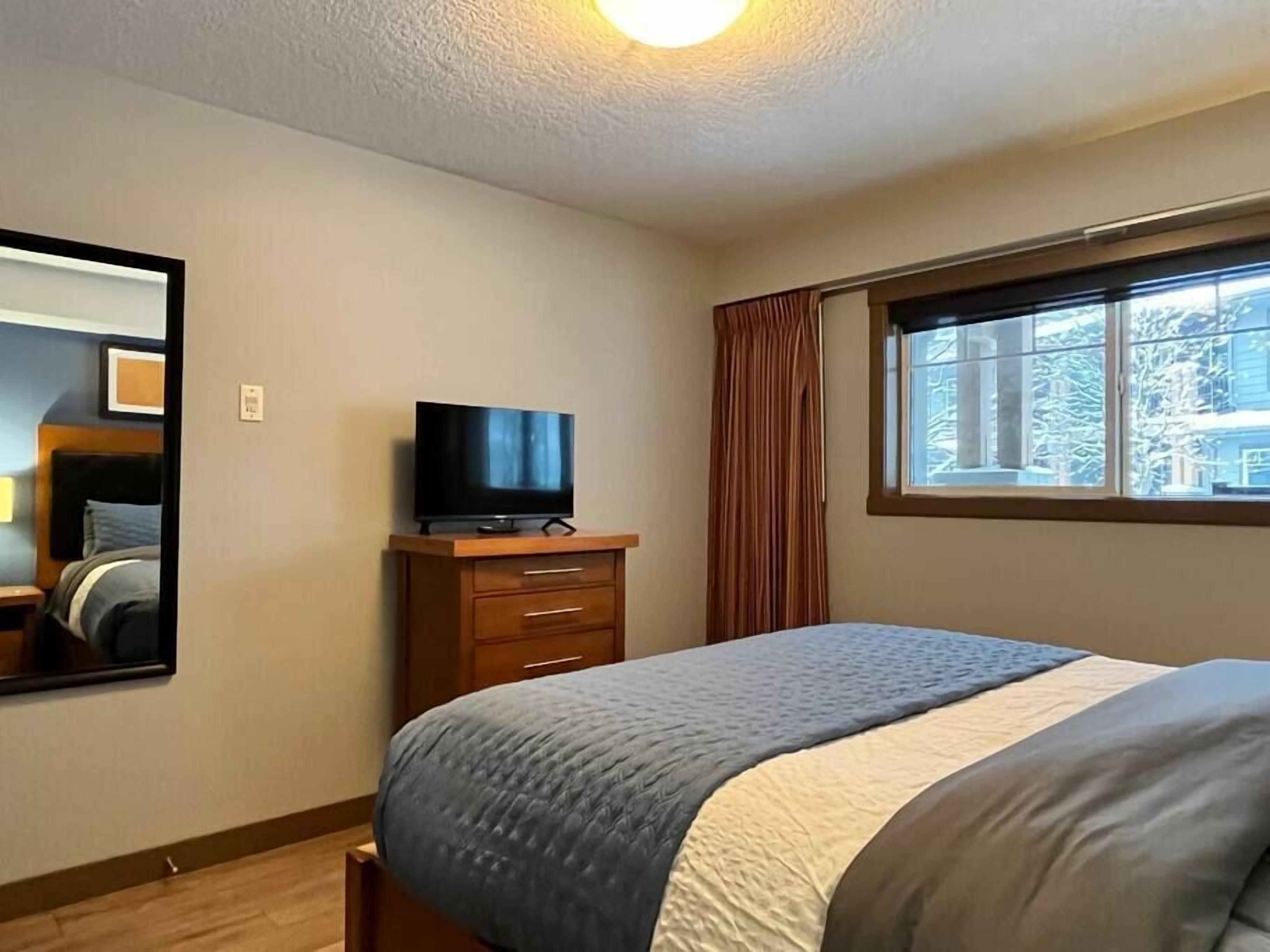 Ein Hotelzimmer mit Bett, Fernseher auf einer Holzkommode, einem Fenster mit Vorhängen und einem Spiegel an der Wand.