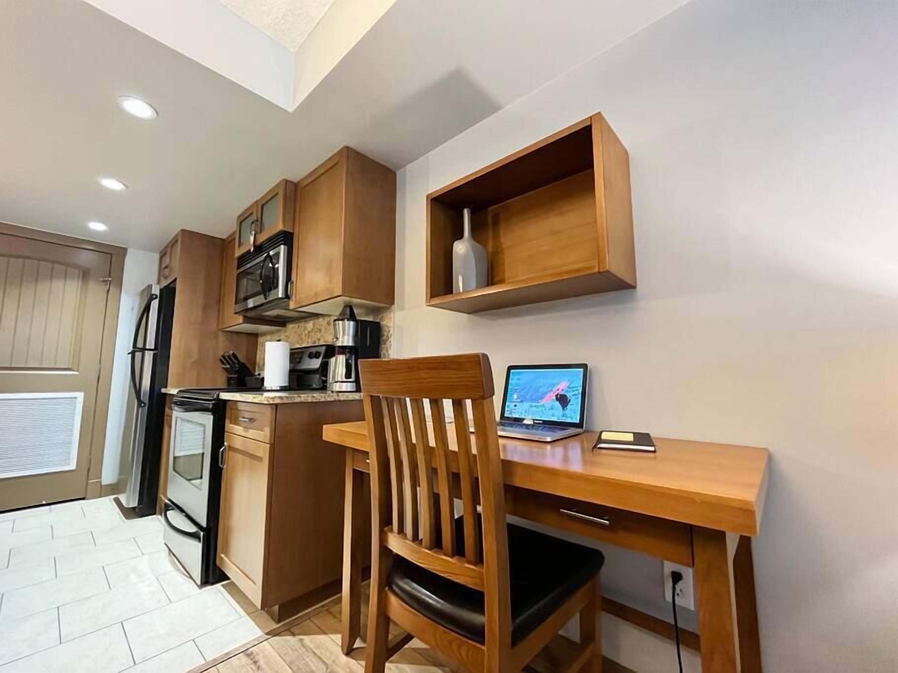 A compact kitchen with wooden cabinets, a microwave, a stove, and a laptop on a desk.