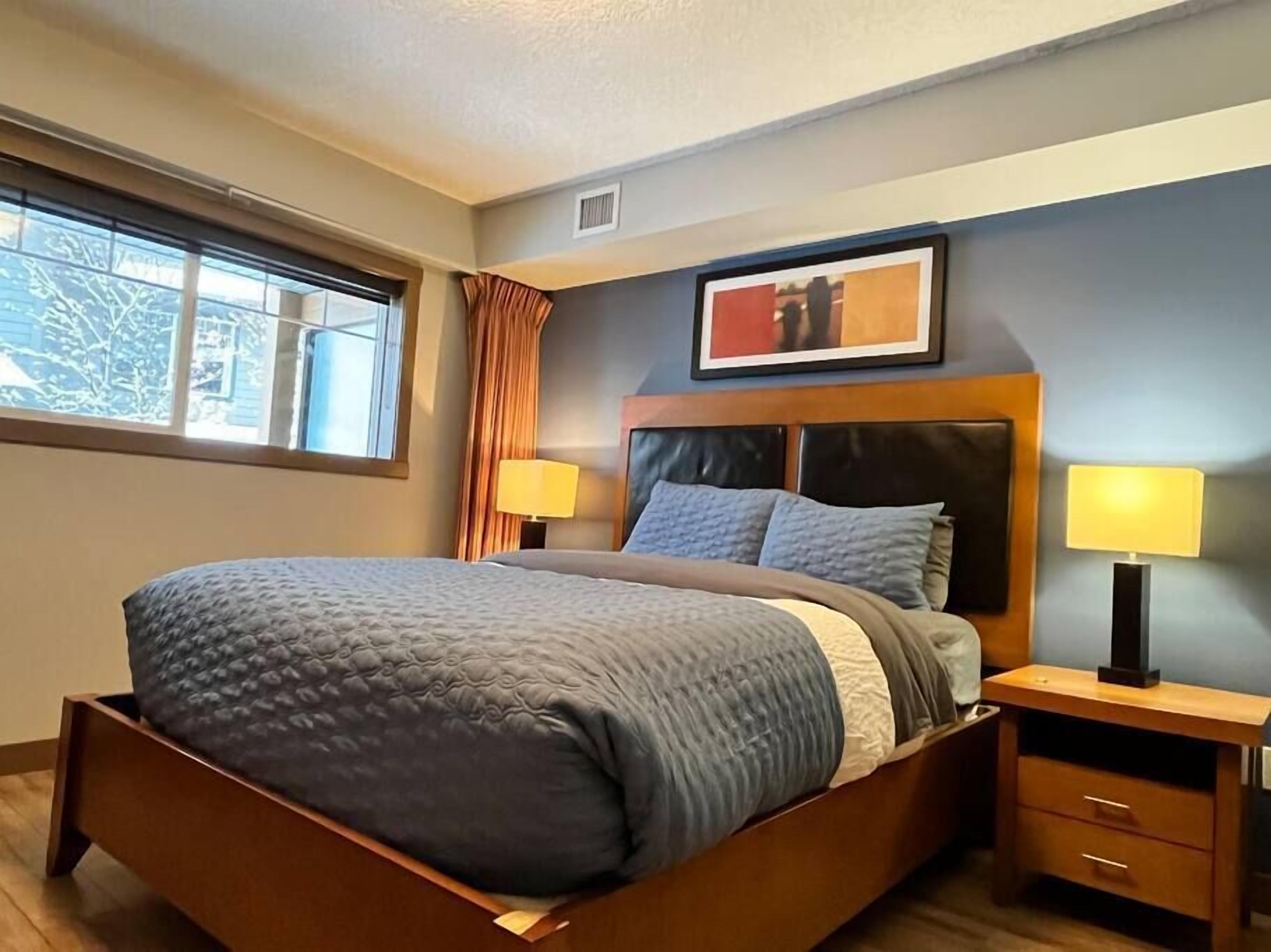 A neatly made bed with a headboard, two bedside tables with lamps, a framed picture above the bed, and a window with curtains.