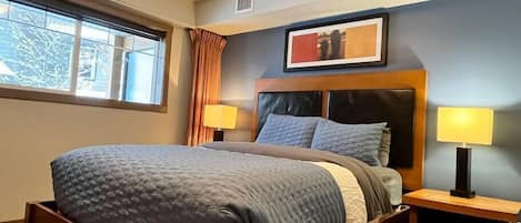 A neatly made bed with a headboard, two bedside tables with lamps, a framed picture above the bed, and a window with curtains.
