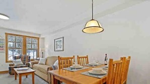 Un coin repas avec une table en bois dressée pour quatre personnes, entourée de chaises en bois. On aperçoit une fenêtre avec des stores, un canapé et une lampe à l'arrière-plan.