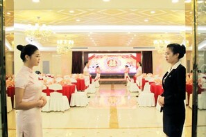Uma sala de banquetes com mesas montadas para um evento formal, um palco com fundo vermelho e um candelabro.