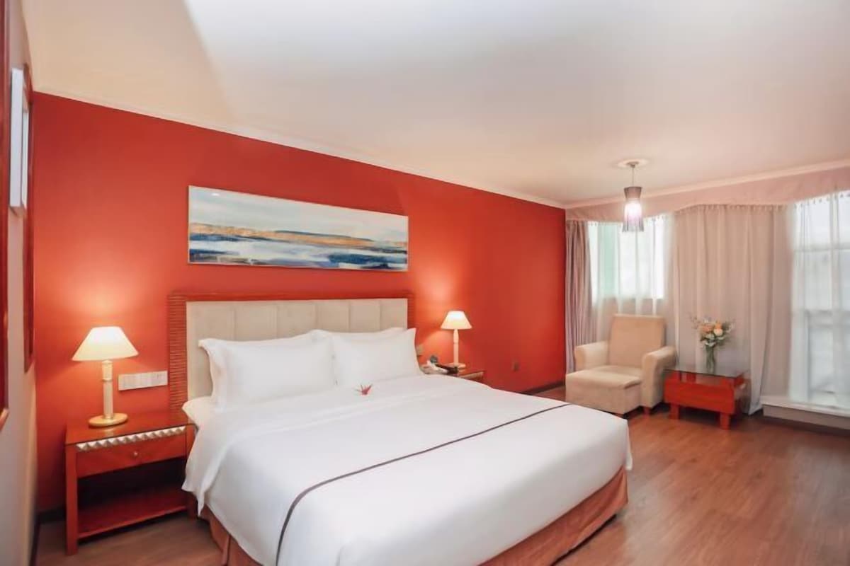A hotel room with a large bed, two bedside tables with lamps, a chair, a window with curtains, and a painting on the wall.