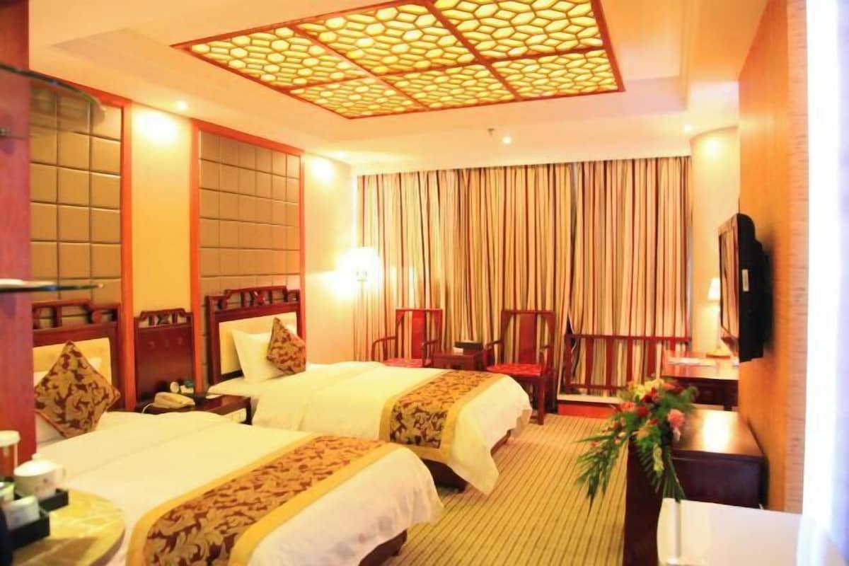 A hotel room with two beds, a large window with curtains, a flat-screen TV mounted on the wall, and a decorative ceiling light fixture.