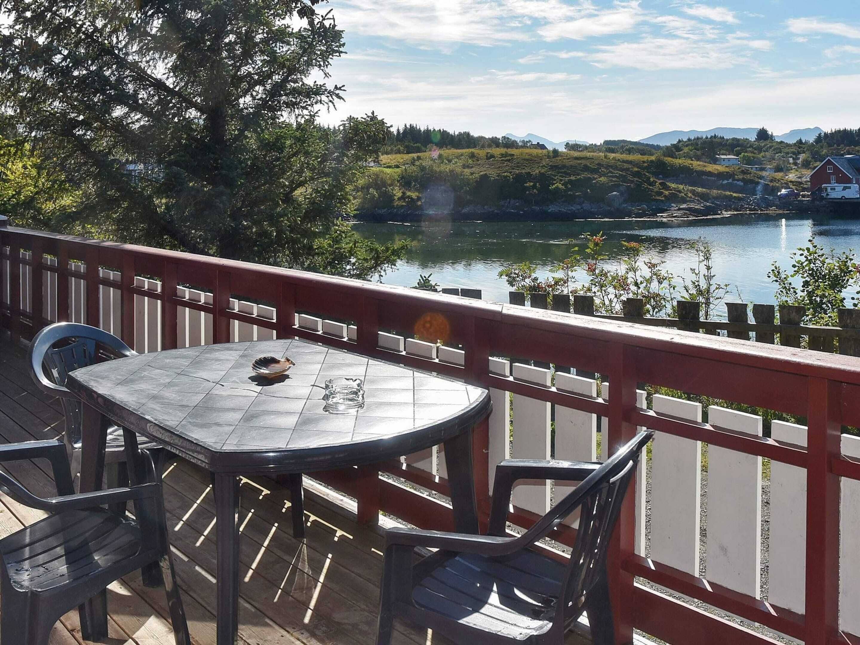 Una terraza con mesa y sillas que da a un lago, con vistas a una casa roja y árboles al fondo.