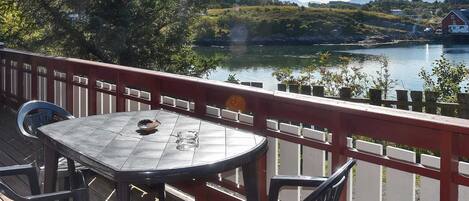Una terraza con mesa y sillas que da a un lago, con vistas a una casa roja y árboles al fondo.
