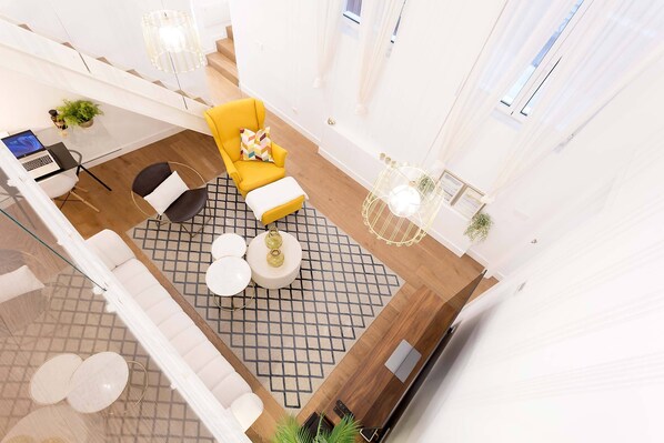 Una sala de estar moderna con un sillón amarillo, un sofá blanco y una alfombra de estampado geométrico. Hay un ordenador portátil sobre una mesita, una planta en maceta y una mesa de centro de cristal.