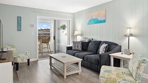 A living room with a grey couch, a coffee table, and a chair with a floral pattern. There is a painting on the wall and a lamp on a side table. - Great Midtown Location with Water Views! (Ocean City)