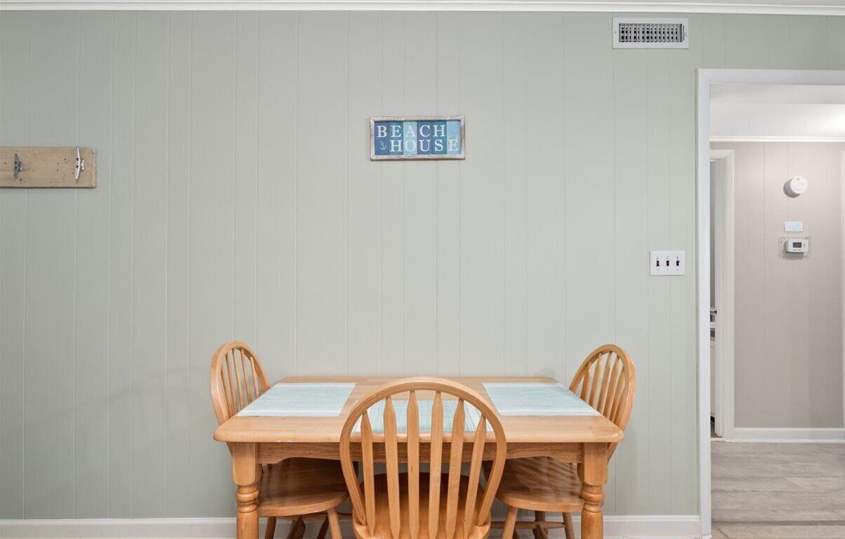 Un comedor con una mesa y sillas de madera, un letrero que dice "BEACH HOUSE" en la pared y una puerta que da a otra habitación.