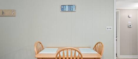 Un comedor con una mesa y sillas de madera, un letrero que dice "BEACH HOUSE" en la pared y una puerta que da a otra habitación.