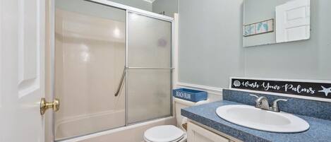 A bathroom with a glass-enclosed shower, a white toilet, a sink with a blue countertop, and a mirror with a 'Wash Your Palms' sign.