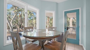A dining area with a round glass table and wooden chairs, large windows with a view of trees, and a teal door. - "Palm Paradise" Golf cart included. (Fripp Island)