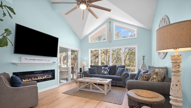 A living room with a fireplace, a ceiling fan, a TV, and large windows offering a view of trees. - "Palm Paradise" Golf cart included. (Fripp Island)